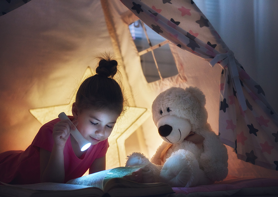 Cute little child is reading a book with flashlights in tent. Happy girl plays at home. Funny lovely kid having fun in children room.