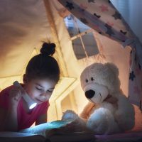 Cute little child is reading a book with flashlights in tent. Happy girl plays at home. Funny lovely kid having fun in children room.